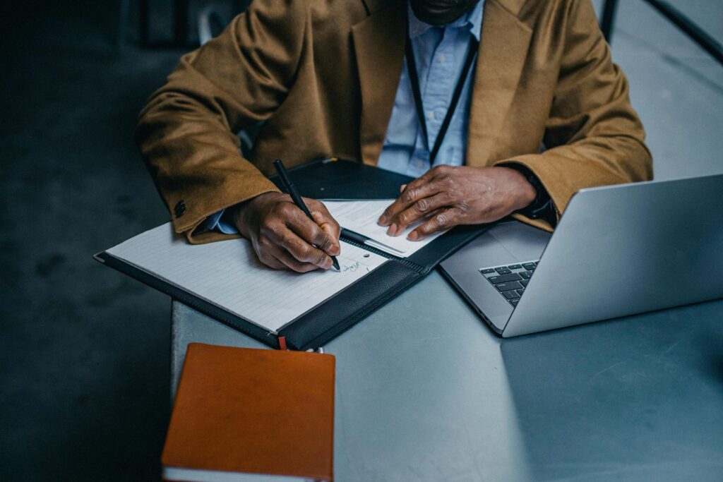 An executive writing in a notebook with a laptop at a stylish indoor workspace.